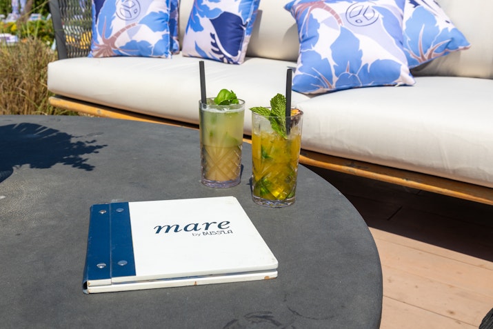 Table with two drinks, a menu labeled "mare," and seating with patterned cushions.
