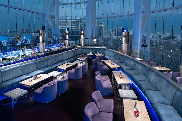 Millennium Al Rawdah Hotel The seating features plush purple chairs and tables, illuminated by blue lighting, creating an inviting and sophisticated atmosphere.
