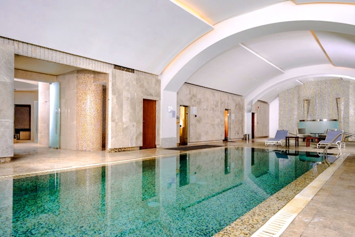 An indoor swimming pool area featuring smooth marble walls and a sleek design. The pool is surrounded by relaxing lounge chairs and soft lighting, creating a tranquil atmosphere.