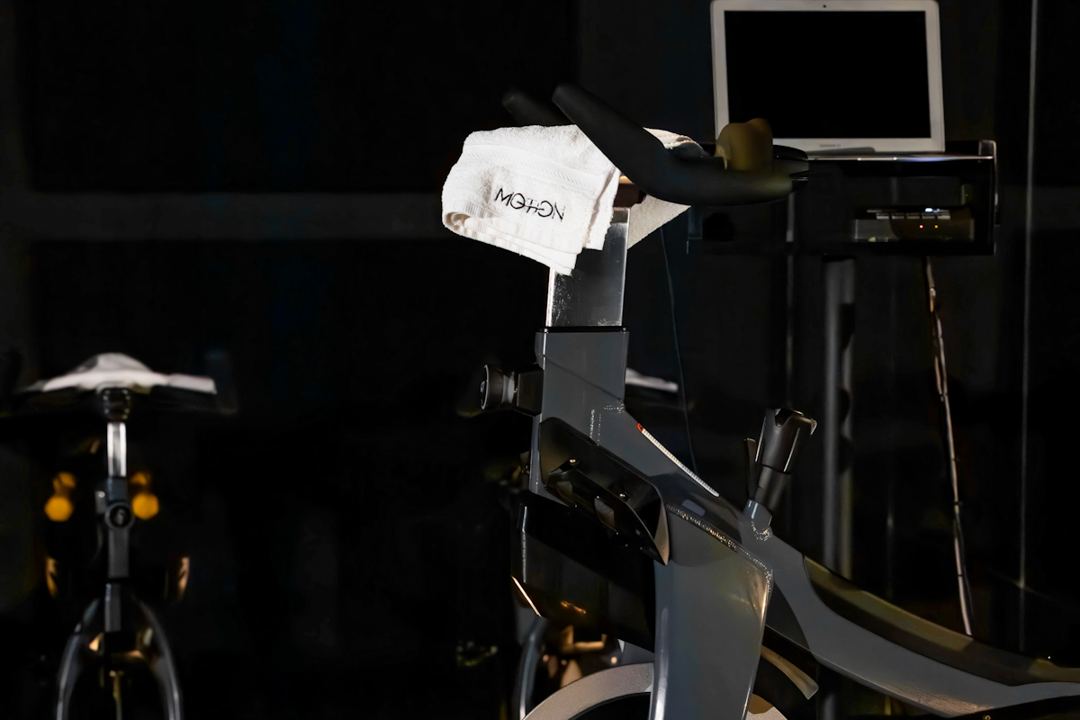 MOTION Cycling A stationary spin bike set up in a dimly lit indoor cycling studio, with a white towel draped over the handlebars. A screen or laptop stands in the background, suggesting a guided workout session. The scene is minimalist and focused on the equipment, ideal for an intense indoor cycling class