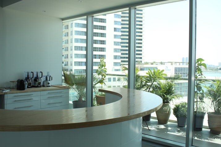 A stylish coffee corner featuring a curved wooden counter in a bright, modern space with floor-to-ceiling windows. Green potted plants line the windows, creating a refreshing atmosphere. The cityscape and clear sky visible through the windows enhance the serene, relaxing ambiance of the corner.