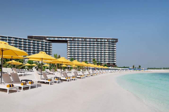 Beach loungers with yellow umbrellas along a white sandy shore, with a modern hotel in the background under a clear blue sky.