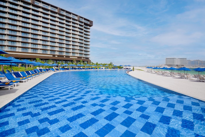 A view of a resort pool with a patterned blue tile design, surrounded by lounge chairs and a tall building in the background under a bright blue sky.