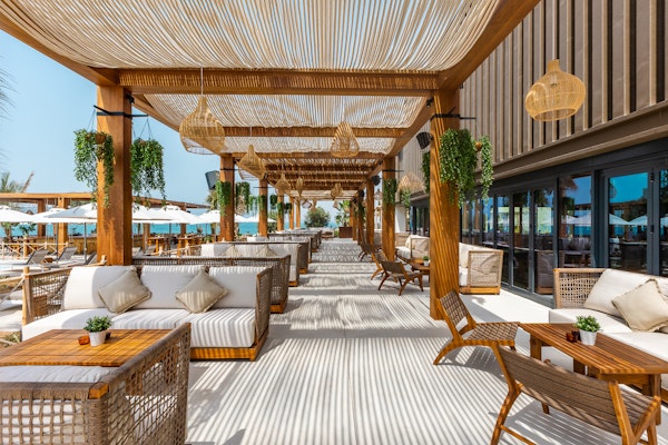 A stylish outdoor dining area with wooden furniture, comfortable seating, and lush greenery, creating a relaxed atmosphere by the beach.