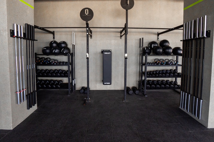 A neatly organized strength training area featuring racks of dumbbells, kettlebells, and medicine balls along with a variety of weightlifting bars mounted on the wall. Two lifting stations with pull-up bars are centered, creating a clean and structured workout space in a modern gym setting