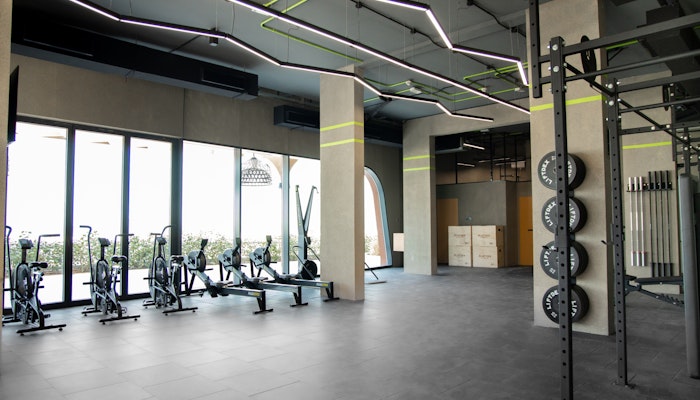 Qudrah Fitness, Sharjah A bright, spacious gym area featuring cardio equipment, such as stationary bikes, near large windows allowing natural light. The ceiling has creative, angular lighting fixtures, and weight plates are organized on racks near the open workout space. The design is minimalist and modern.