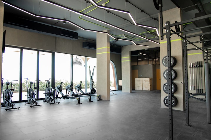A bright, spacious gym area featuring cardio equipment, such as stationary bikes, near large windows allowing natural light. The ceiling has creative, angular lighting fixtures, and weight plates are organized on racks near the open workout space. The design is minimalist and modern.