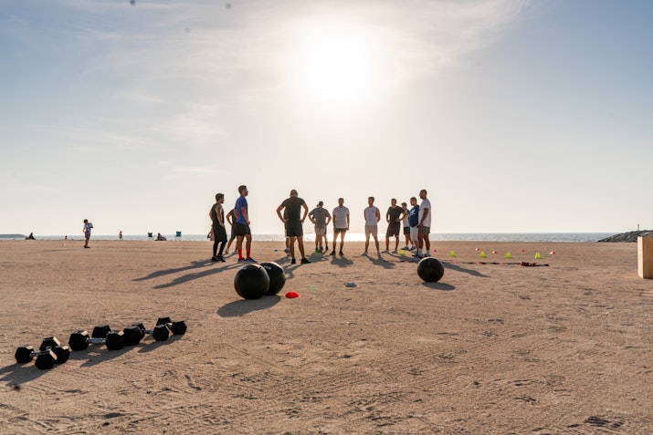 A group of people gathered on the beach for a fitness bootcamp session, with the sun rising in the background. Exercise equipment, such as medicine balls and kettlebells, is spread out on the sand. The scene captures an early morning workout with a serene beach setting