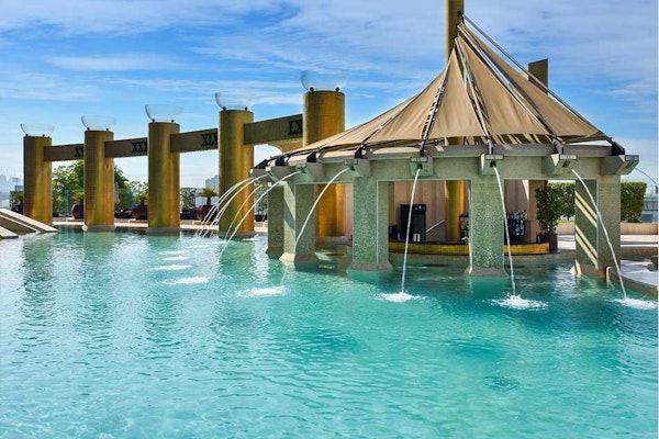 Resort-style pool with a thatched pavilion and water fountains, surrounded by tall columns and blue sky