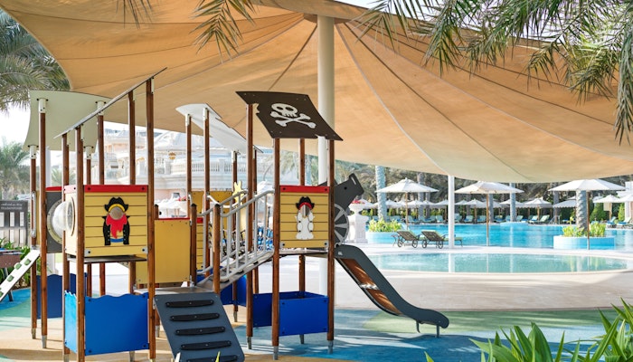 Raffles The Palm Dubai A pirate theme, complete with a slide and climbing structures, shaded by a large canopy, located near a swimming pool area.