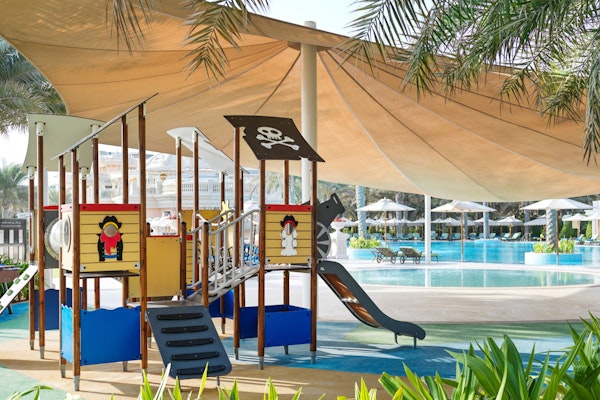 A pirate theme, complete with a slide and climbing structures, shaded by a large canopy, located near a swimming pool area.