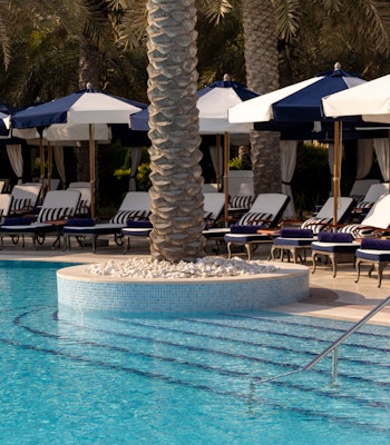 Raffles The Palm Dubai A serene pool area featuring lounge chairs under blue and white umbrellas, surrounded by palm trees.