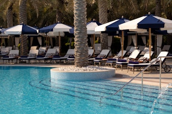 A serene pool area featuring lounge chairs under blue and white umbrellas, surrounded by palm trees.