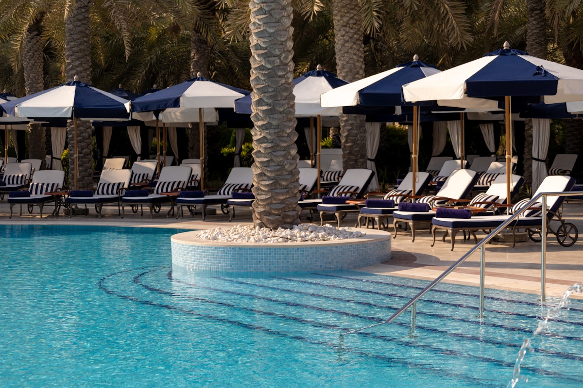 A serene pool area featuring lounge chairs under blue and white umbrellas, surrounded by palm trees.