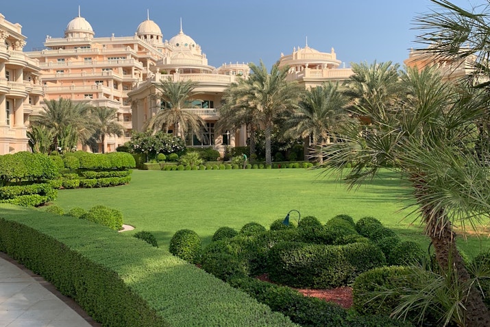 Garden with lush green lawns and neatly trimmed hedges, surrounded by an elegant hotel building characterized by domes and architectural detail.