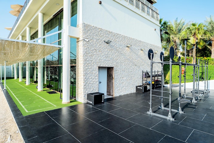 Open-air fitness space featuring weightlifting racks on black flooring, adjacent to a turf walkway and modern building with glass walls.