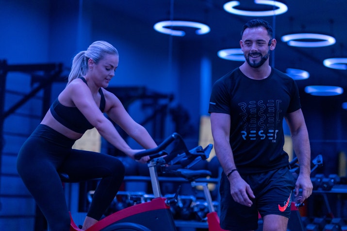 Reset Fitness A woman exercising on a stationary bike while a man stands beside her, both in a gym with blue lighting.