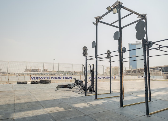 Outdoor gym setup featuring tall metal pull-up bars, SkiErg machines, and large tractor tires on a paved area, with a distant view of city buildings and a sign that reads 'Now it’s your turn