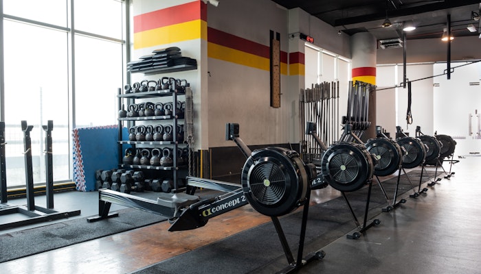 SK Fitness Gym area featuring multiple rowing machines aligned in a row, next to a weight rack with kettlebells, barbells, and other gym equipment, set against a backdrop with large windows and a colorful wall stripe design.