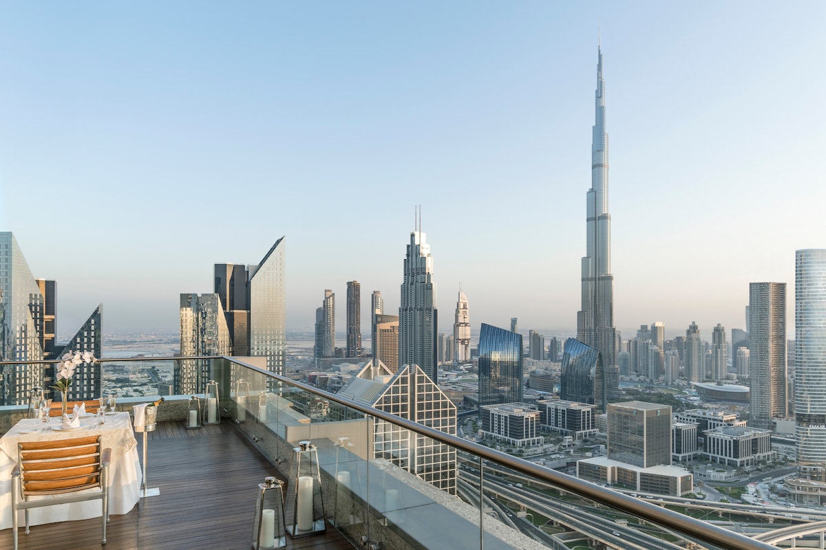 A rooftop dining area with a table set for two, overlooking a panoramic view of the Dubai skyline, featuring the iconic Burj Khalifa.