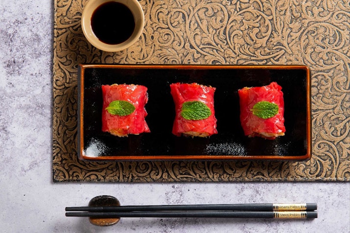Shangri-La Dubai Three pieces of sushi topped with slices of fish and garnished with mint leaves, served on a black plate with soy sauce in a small bowl and chopsticks.