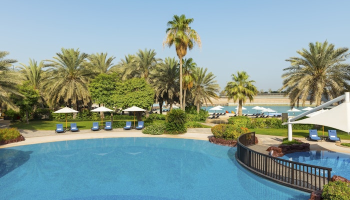 Sheraton Abu Dhabi Hotel & Resort A tranquil resort pool area surrounded by lush greenery and palm trees, with sun loungers arranged neatly around the pool, creating an inviting atmosphere for relaxation.