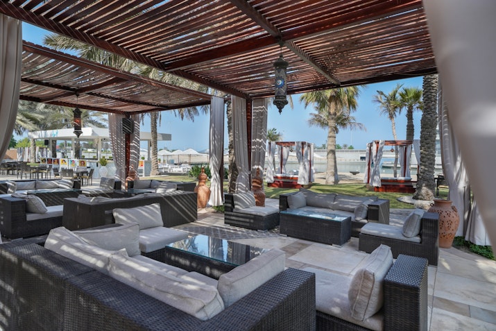 A stylish outdoor lounge area featuring dark wicker furniture with plush cushions, shaded by a wooden canopy.