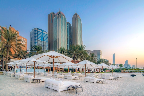 A serene beach scene featuring white lounge chairs and umbrellas on soft sand, with tall palm trees in the foreground and modern skyscrapers in the background, all under a soft sunset sky.