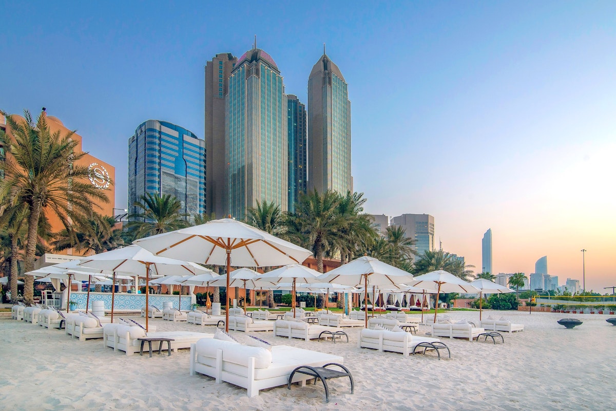 A serene beach scene featuring white lounge chairs and umbrellas on soft sand, with tall palm trees in the foreground and modern skyscrapers in the background, all under a soft sunset sky.