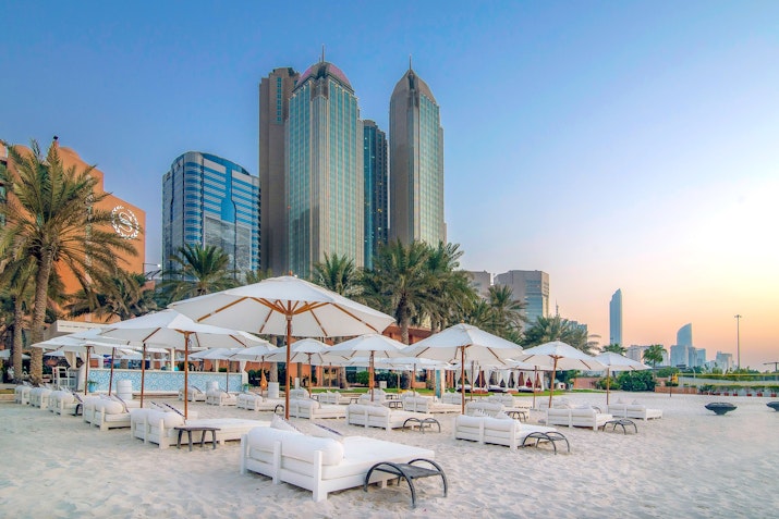 A serene beach scene featuring white lounge chairs and umbrellas on soft sand, with tall palm trees in the foreground and modern skyscrapers in the background, all under a soft sunset sky.
