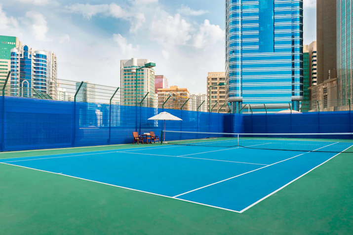 A bright blue tennis court in an urban setting, framed by tall city buildings and secured with blue fencing, offering a modern and inviting space for tennis enthusiasts.