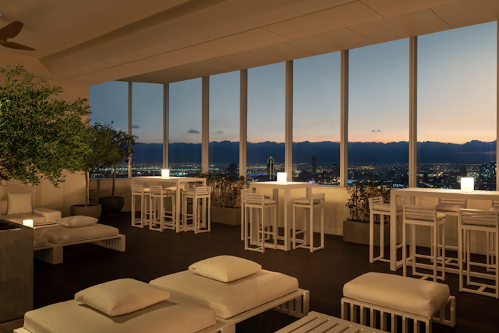A chic lounge area with white furniture and a view of a city skyline at sunset, featuring large windows that allow natural light to fill the space, complemented by plants and elegantly arranged tables.