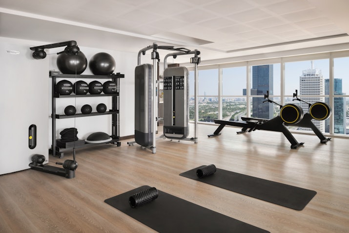 A modern gym featuring exercise equipment including weights, a multi-functional machine, and yoga mats, with large windows offering a view of the city skyline.
