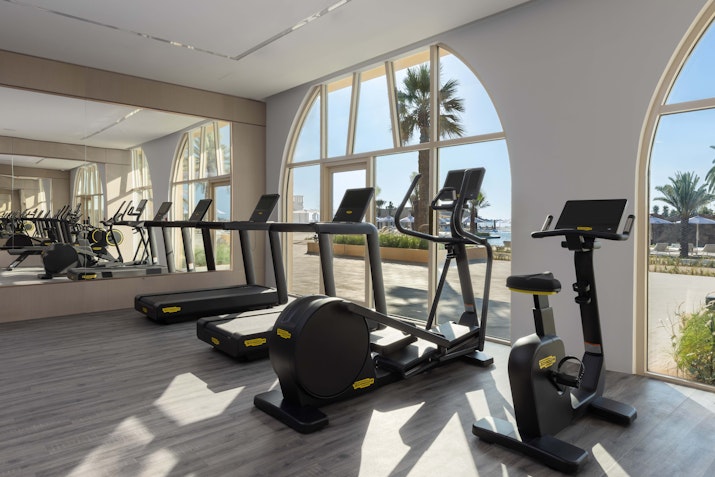 A well-lit gym featuring modern fitness equipment like treadmills and stationary bikes, with large windows offering a view of palm trees outside.