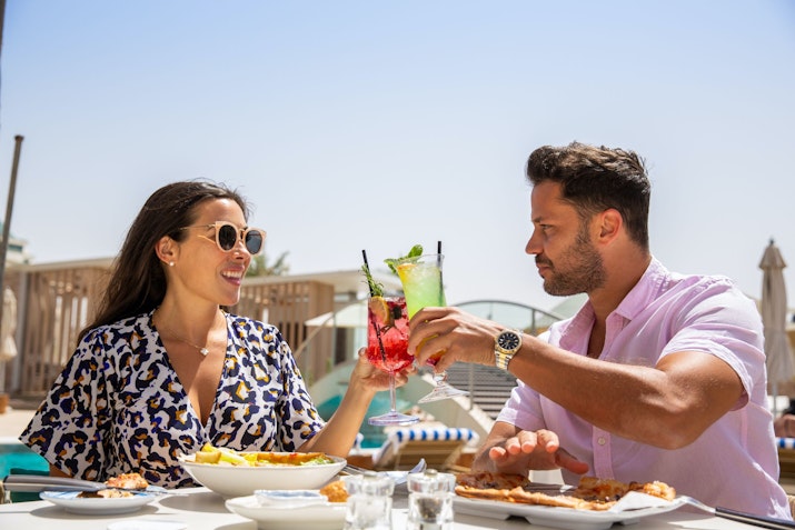 A couple enjoying drinks and a meal outdoors, with a sunny beach in the background