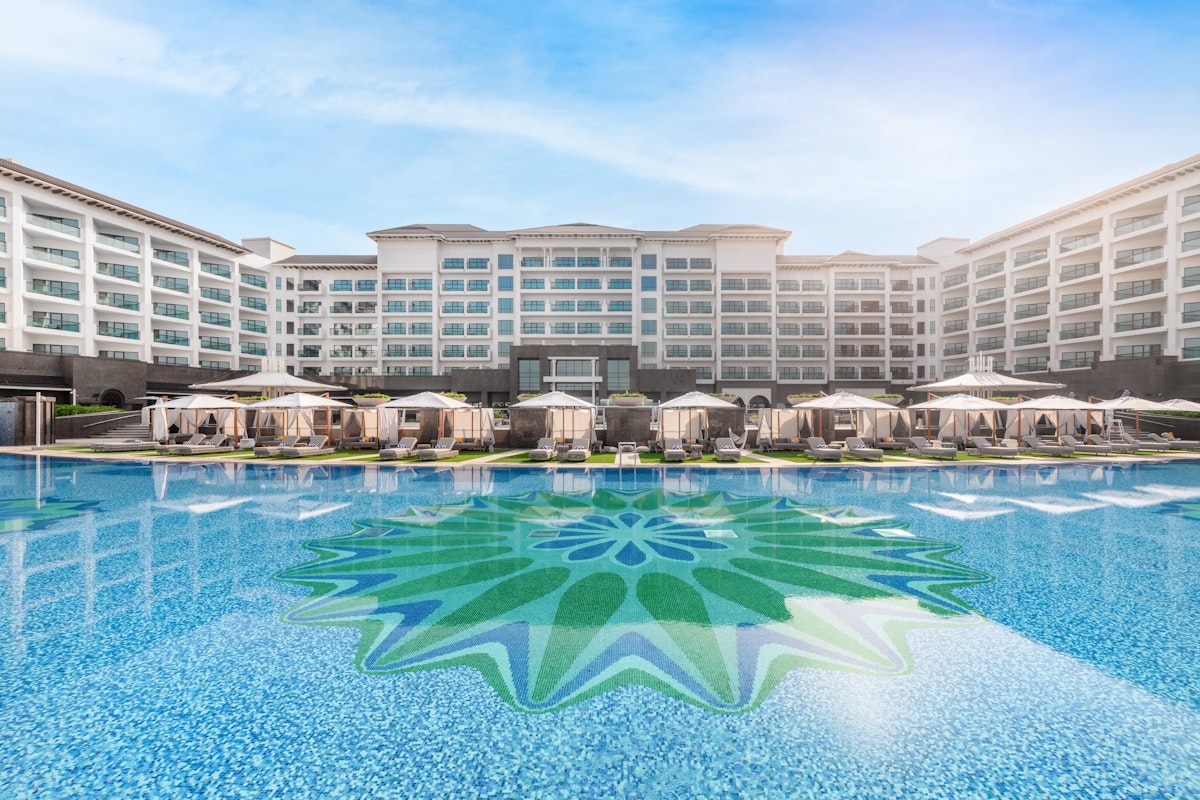 A luxury hotel with a large swimming pool featuring a decorative mosaic design, surrounded by cabanas and lounge chairs, under a bright blue sky.