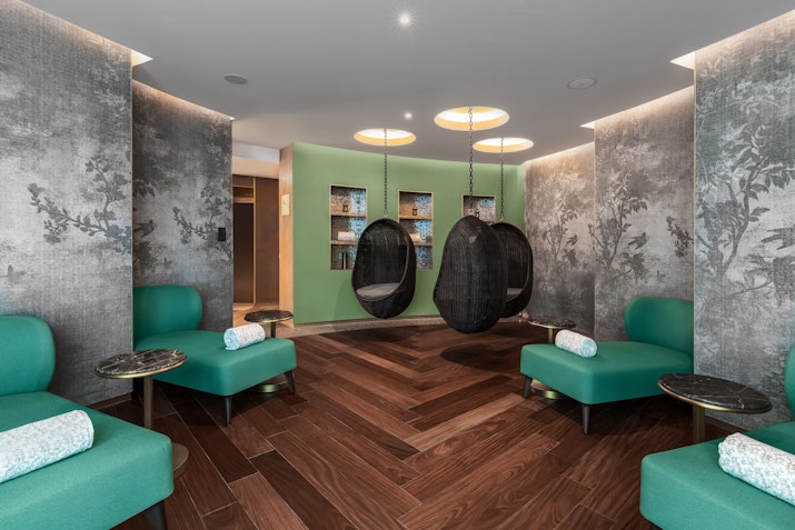 A stylish relaxation area with green seating, circular hanging chairs, and soft lighting, set against textured walls.