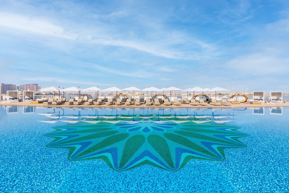A swimming pool with a vibrant mosaic design at the center, lined with cabanas and lounge chairs, set against a bright blue sky.