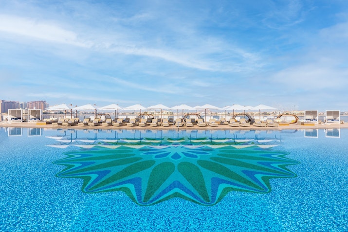 A swimming pool with a vibrant mosaic design at the center, lined with cabanas and lounge chairs, set against a bright blue sky.