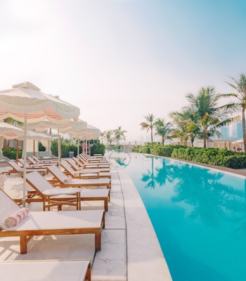 The 305 Dubai A serene poolside scene featuring lounge chairs with colorful towels, elegant cabanas, and palm trees, set against a backdrop of modern buildings and a clear sky.