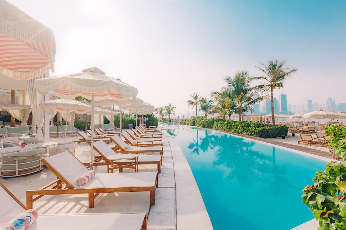 A serene poolside scene featuring lounge chairs with colorful towels, elegant cabanas, and palm trees, set against a backdrop of modern buildings and a clear sky.