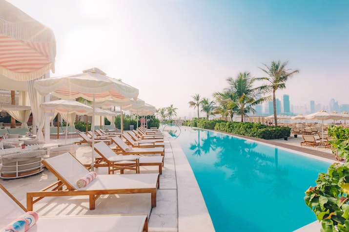 A serene poolside scene featuring lounge chairs with colorful towels, elegant cabanas, and palm trees, set against a backdrop of modern buildings and a clear sky.