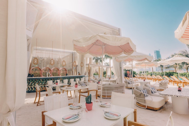 The 305 Dubai An outdoor dining area featuring white tables and chairs, bright sunshades, and a relaxed ambiance, with a glimpse of a pool and lush surroundings in the background.