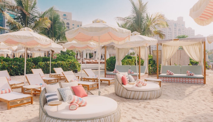 The 305 Dubai A cozy beach lounge area featuring circular daybeds and sun loungers with vibrant pillows, surrounded by palm trees and sunshades, creating a tropical and relaxing atmosphere.