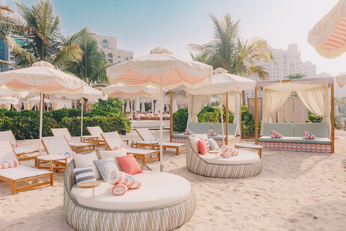 The 305 Dubai A cozy beach lounge area featuring circular daybeds and sun loungers with vibrant pillows, surrounded by palm trees and sunshades, creating a tropical and relaxing atmosphere.