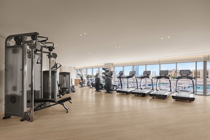 Well-lit gym with treadmills, exercise machines, and large windows offering scenic city views, providing a spacious and airy workout environment.