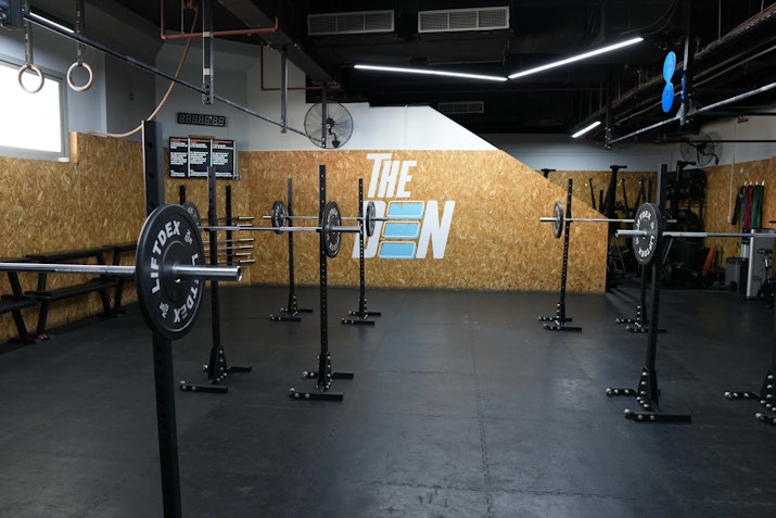 A spacious gym setup at The Den featuring squat racks and weightlifting equipment against a backdrop with The Den logo, designed for strength and conditioning training.
