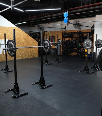 The Den A spacious gym setup at The Den featuring squat racks and weightlifting equipment against a backdrop with The Den logo, designed for strength and conditioning training.