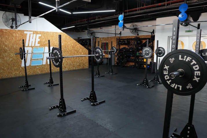 A spacious gym setup at The Den featuring squat racks and weightlifting equipment against a backdrop with The Den logo, designed for strength and conditioning training.