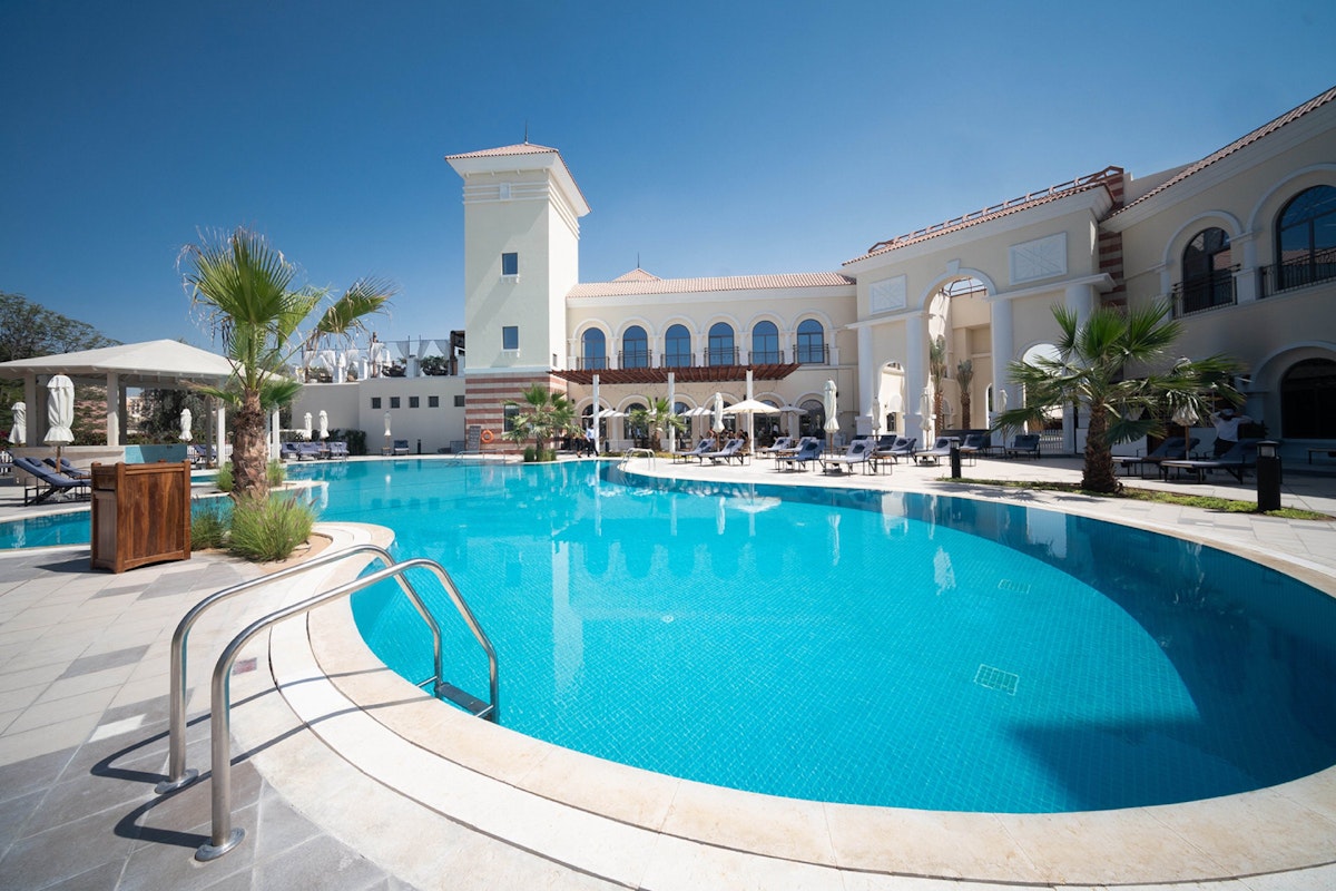 Swimming pool area at a resort, featuring a clear blue pool with a curved edge, surrounded by palm trees and sun loungers.
